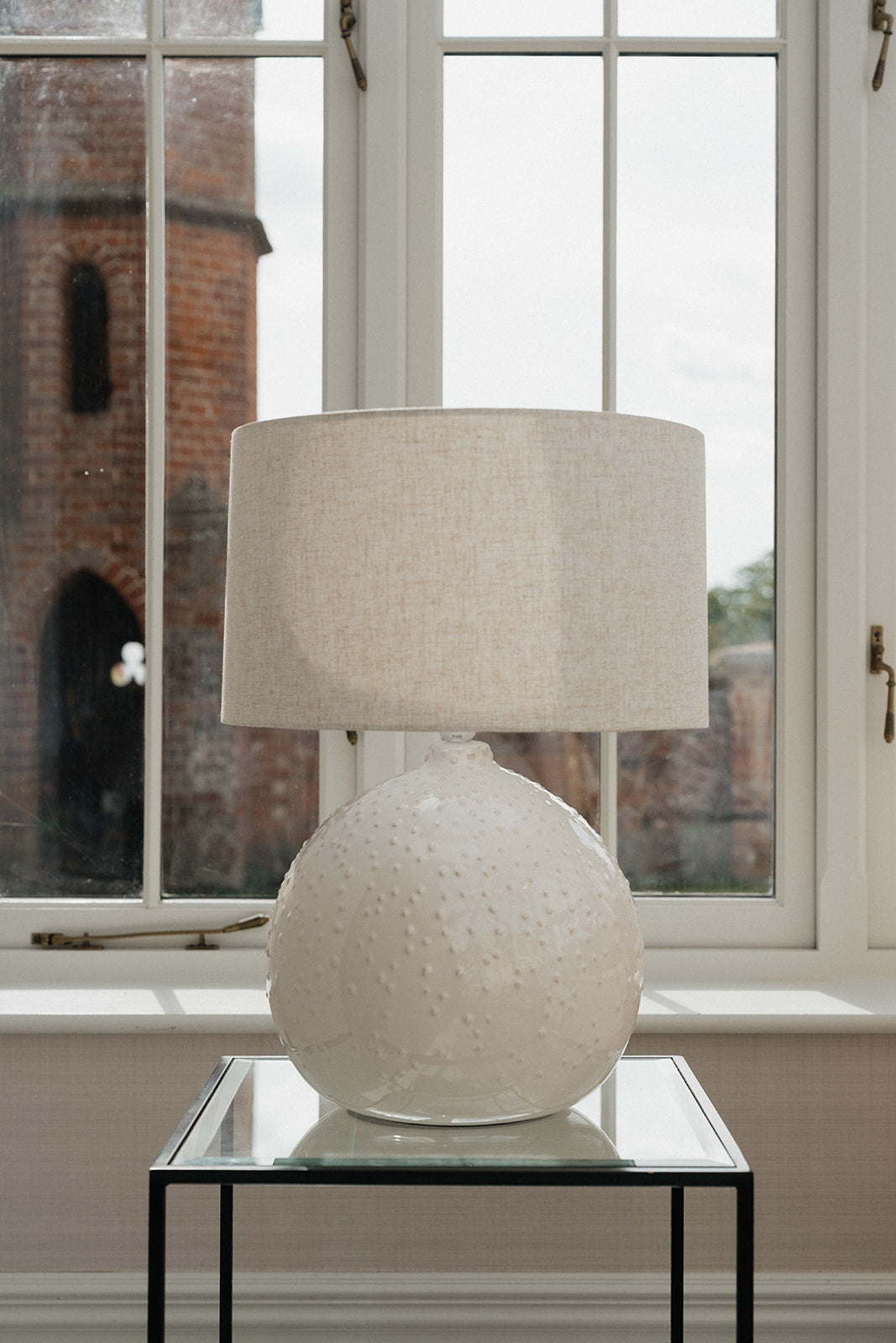 A table lamp with a textured white ceramic globe base and a beige linen drum shade, displayed on a glass-topped side table in front of a large window.