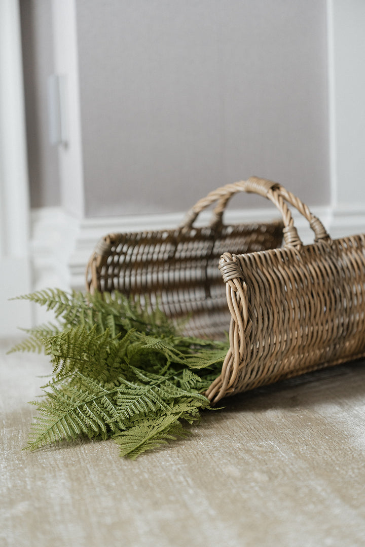 A natural woven wicker basket with two handles lies on a light beige floor, with green fern fronds resting in front of it against a softly blurred gray wall background.