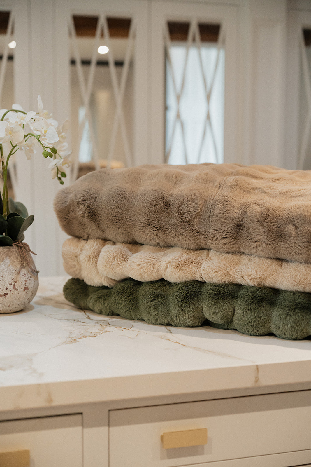 Three plush faux fur throw blankets in taupe, cream, and sage green, stacked neatly on a white marble countertop with a white orchid in a vase to the left.