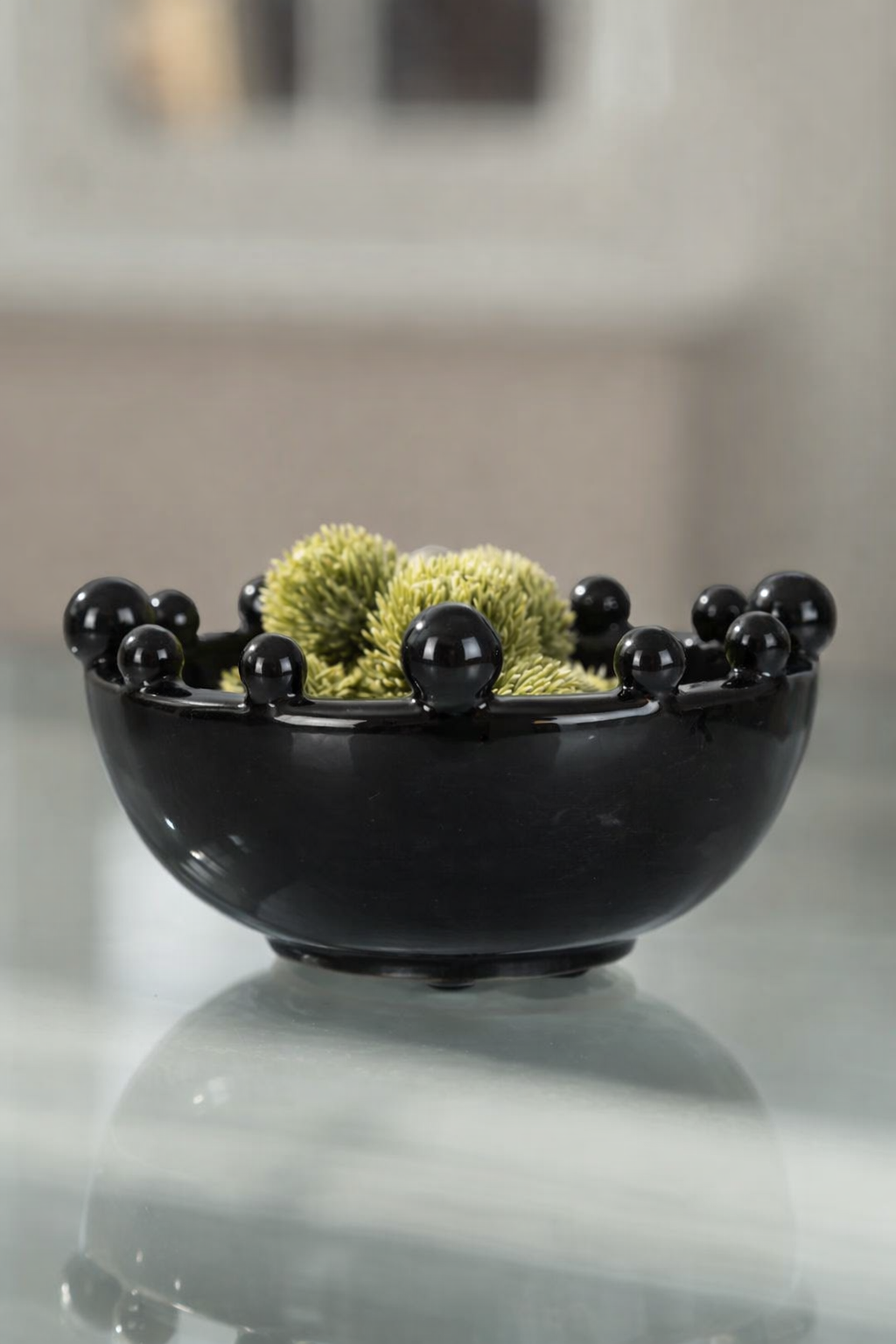 Black decorative bowl with spherical elements and green moss-like material on a reflective surface.