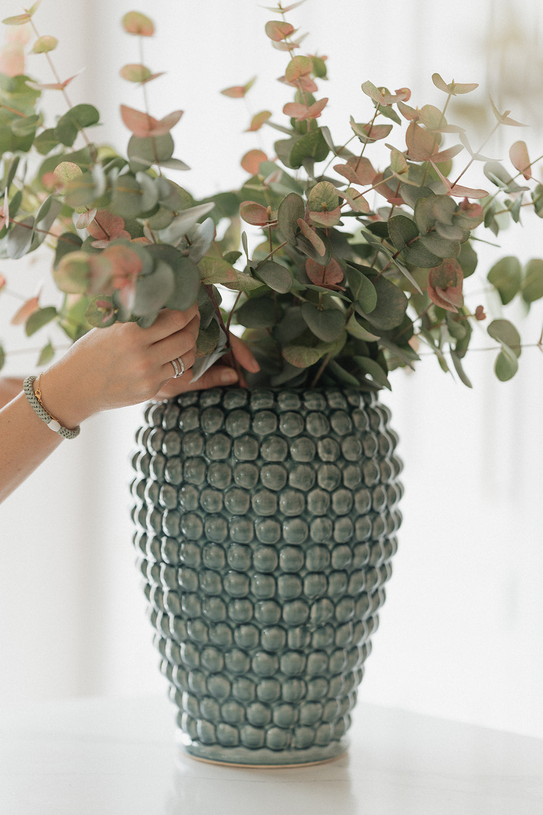 A textured ceramic vase with a blue-green glazed finish, filled with eucalyptus branches, with a person's hand adjusting the foliage.