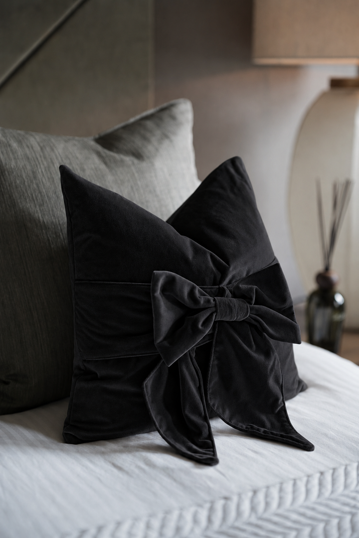 A black velvet decorative throw pillow with a large bow detail, styled on a white textured bed alongside a gray pillow in a softly lit bedroom setting.