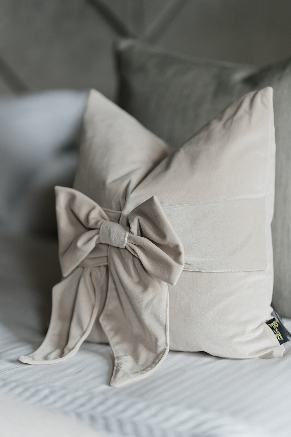 A light beige decorative throw pillow with a large fabric bow detail on the front, styled on a bed with gray pillows in the background.