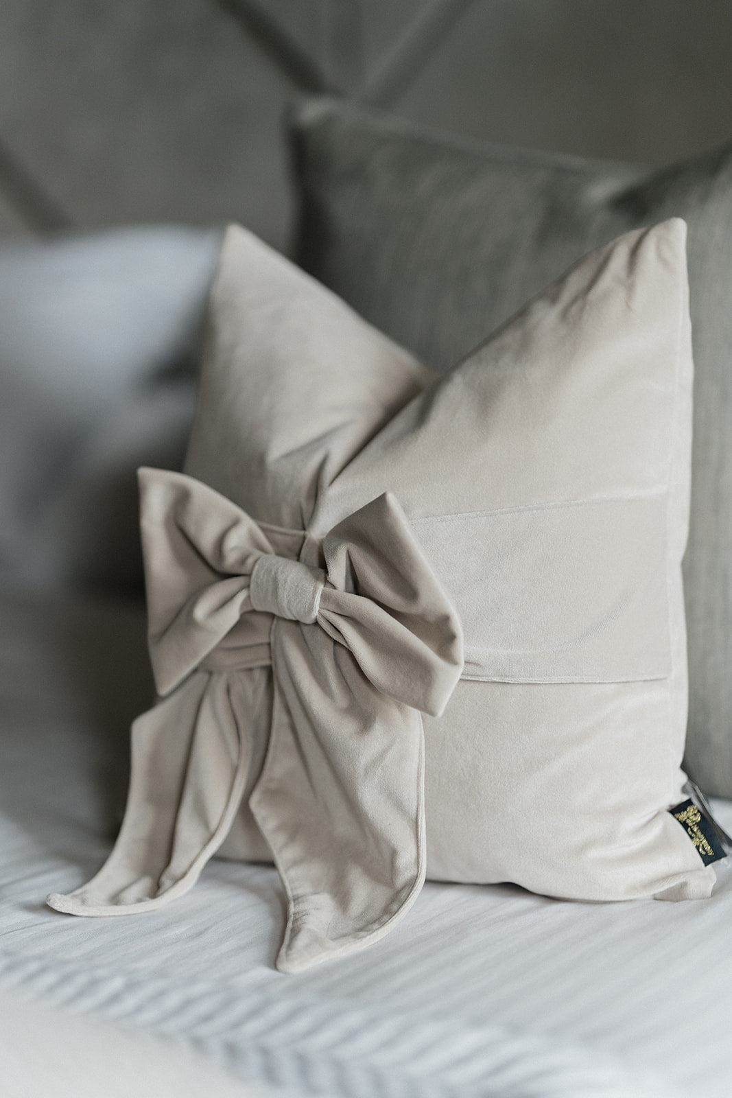 A light beige decorative throw pillow with a large fabric bow detail on the front, styled on a bed with gray pillows in the background.