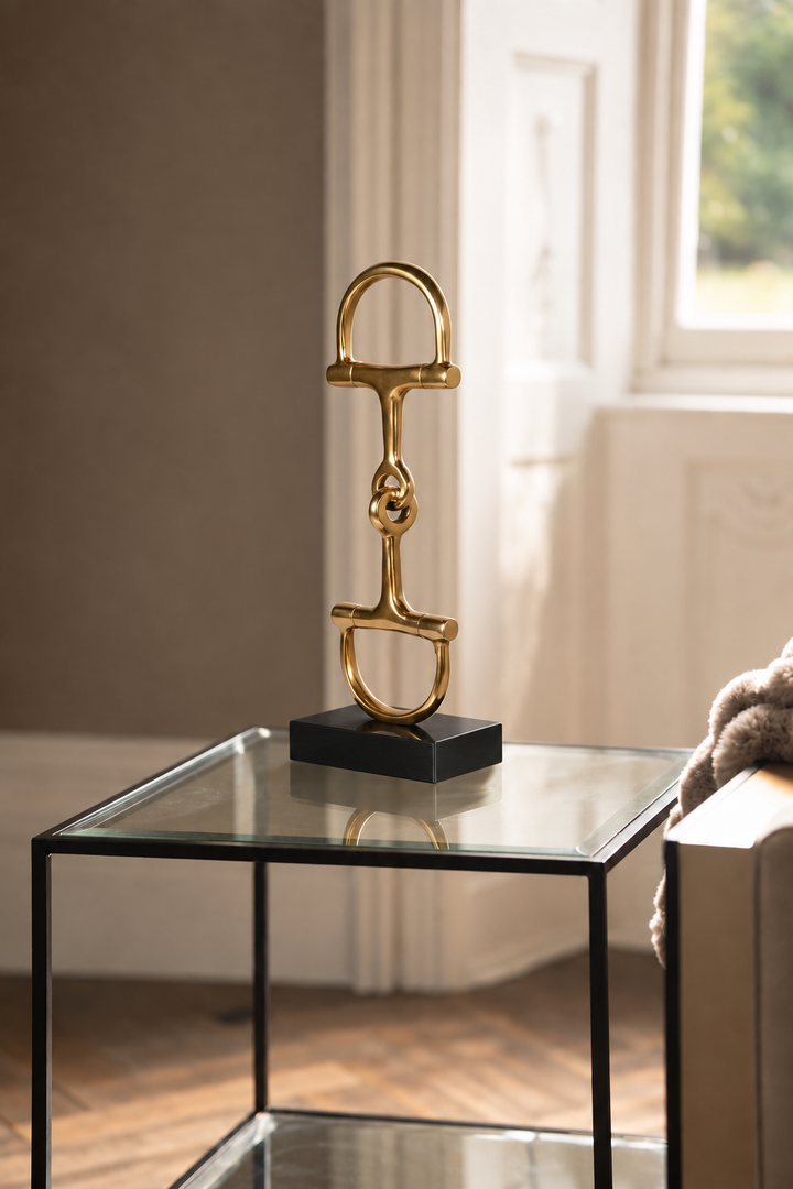 A gold horse bit sculpture on a black rectangular base, displayed on a modern glass-top side table in a softly lit room.
