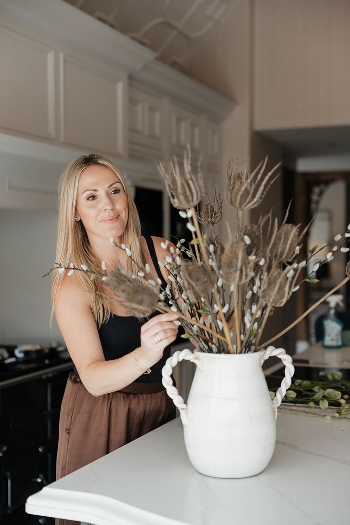 A woman in a kitchen setting holds a large dried floral arrangement in a white ceramic pitcher vase with twisted handles.