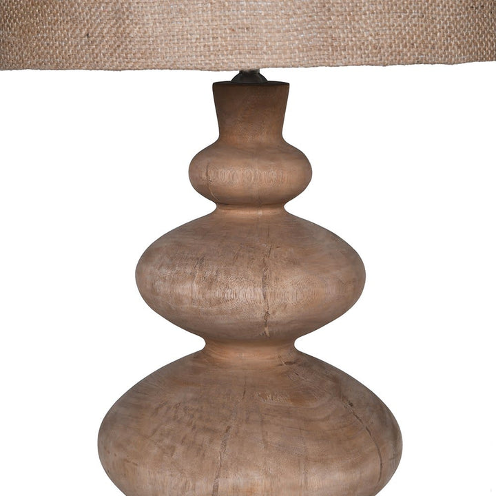 A close-up of a table lamp with a sculptural, multi-tiered wooden base in a natural light brown finish and a woven jute lampshade, shown against a plain white background.