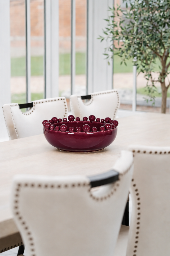 A glossy deep burgundy ceramic bowl filled with dark red cherries, styled on a light wood dining table surrounded by cream upholstered chairs with nailhead trim.