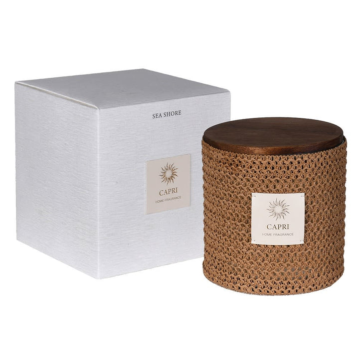 A Capri Sea Shore home fragrance candle in a woven brown jar with a dark wooden lid, shown next to its textured white packaging box.
