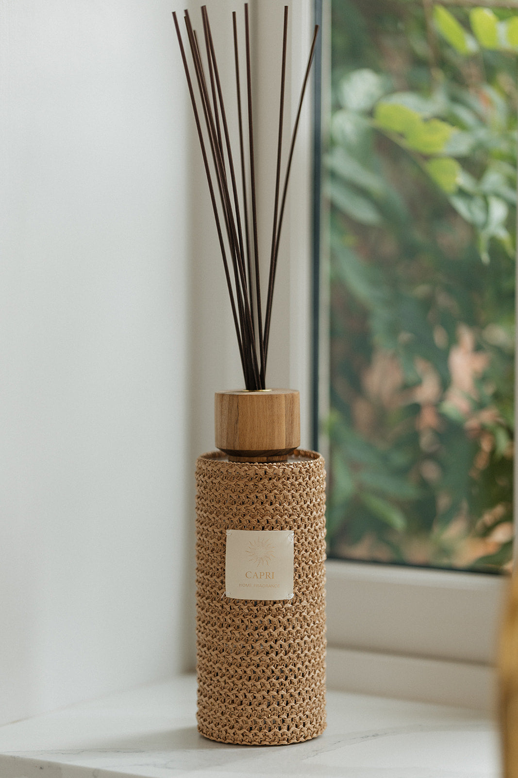 A reed diffuser bottle wrapped in a natural woven straw cover, with dark brown reeds inserted and a small white label on the front, placed on a windowsill with a blurred outdoor garden view in the background.