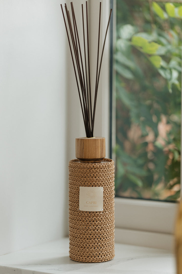 A reed diffuser bottle wrapped in a natural woven straw cover, with dark brown reeds inserted and a small white label on the front, placed on a windowsill with a blurred outdoor garden view in the background.