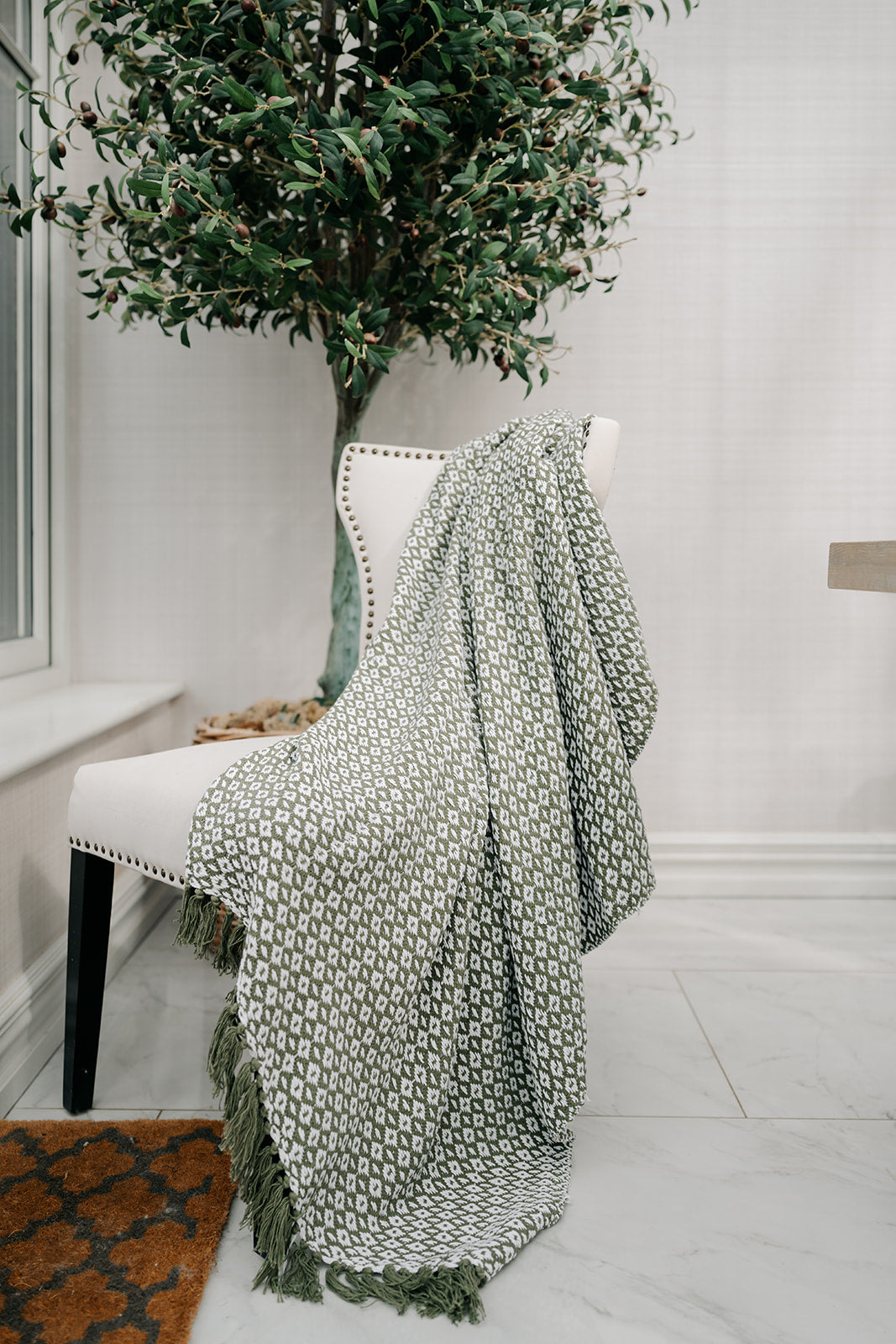 A green and white patterned throw blanket with fringe is draped over a white upholstered chair in a bright, modern room with a large potted tree.