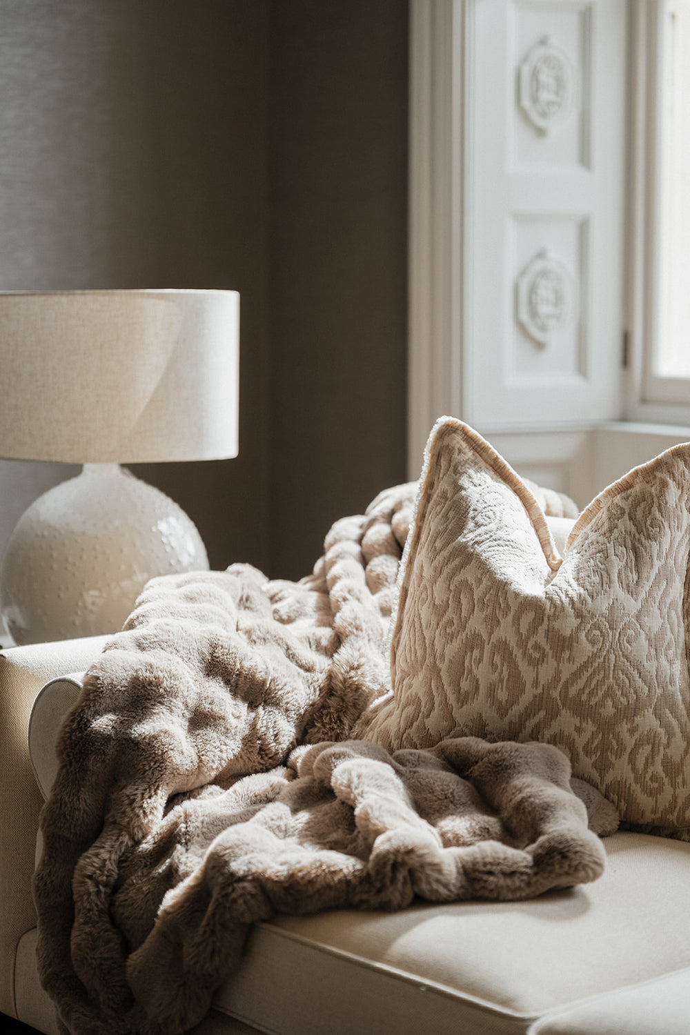 A cozy beige throw blanket and matching textured pillow arranged on a cream sofa in a softly lit room, with a lamp and window visible in the background.