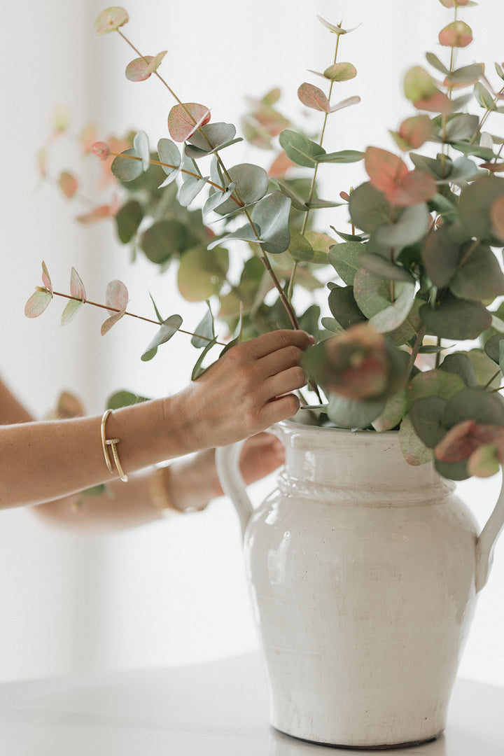 A hand arranging round green eucalyptus leaves in a white ceramic vase with two handles, set against a bright, softly blurred background.