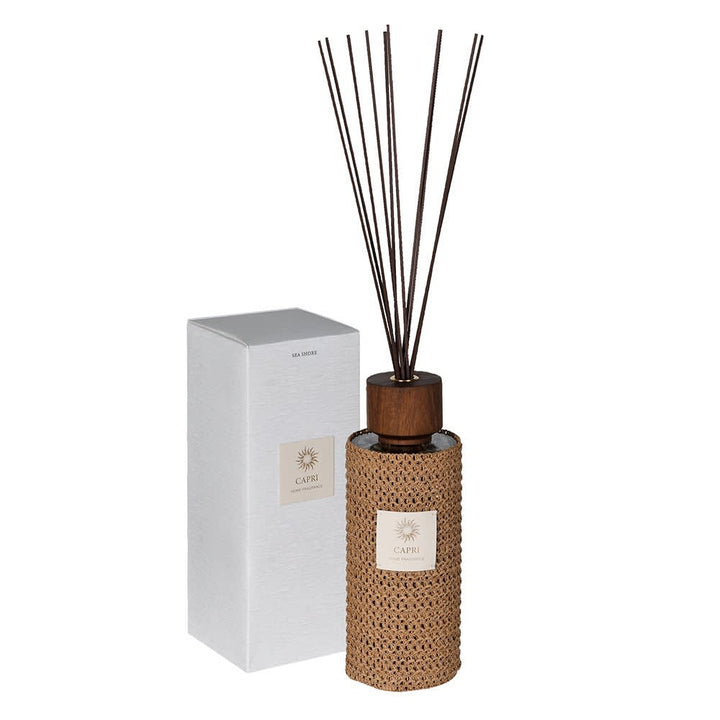 A reed diffuser bottle with a woven tan wrap, dark wood cap, and dark reeds, shown next to its white box on a white background.