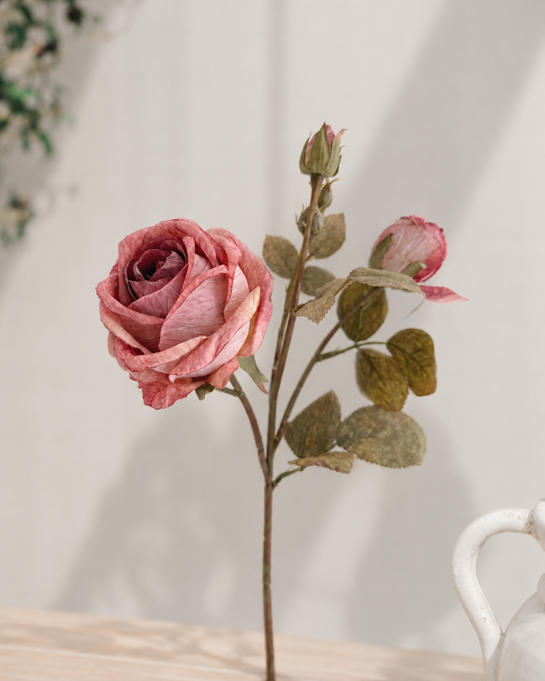 A single artificial pink rose stem with two buds and muted green-brown leaves, displayed on a light wooden surface against a soft off-white background.