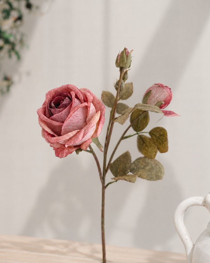 A single artificial pink rose stem with two buds and muted green-brown leaves, displayed on a light wooden surface against a soft off-white background.