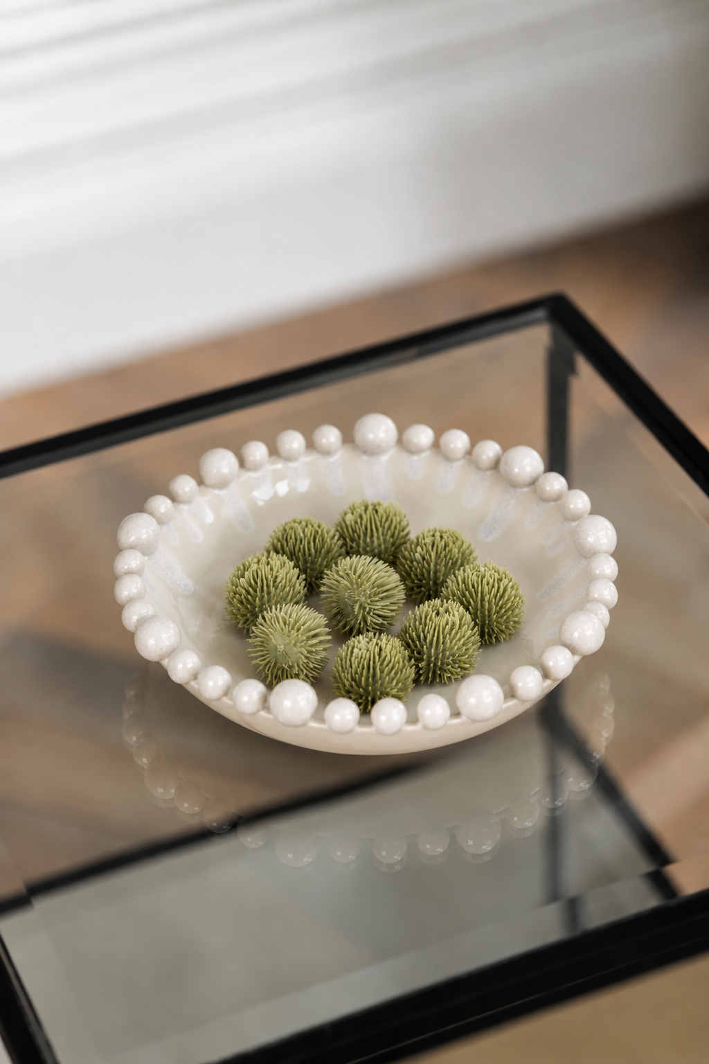 A white ceramic bowl with a beaded border, filled with green decorative pom-pom balls, placed on a glass-top side table.