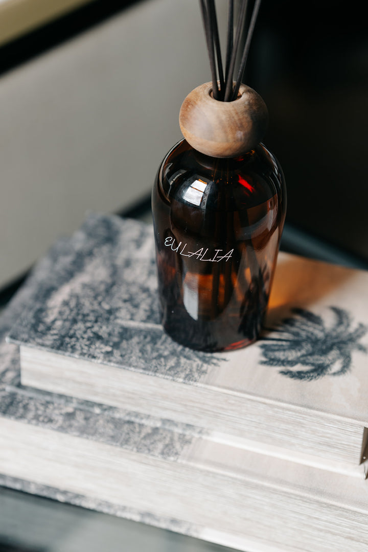 An amber glass reed diffuser bottle with a round wooden top and black reeds, resting on stacked books. The bottle is labeled &