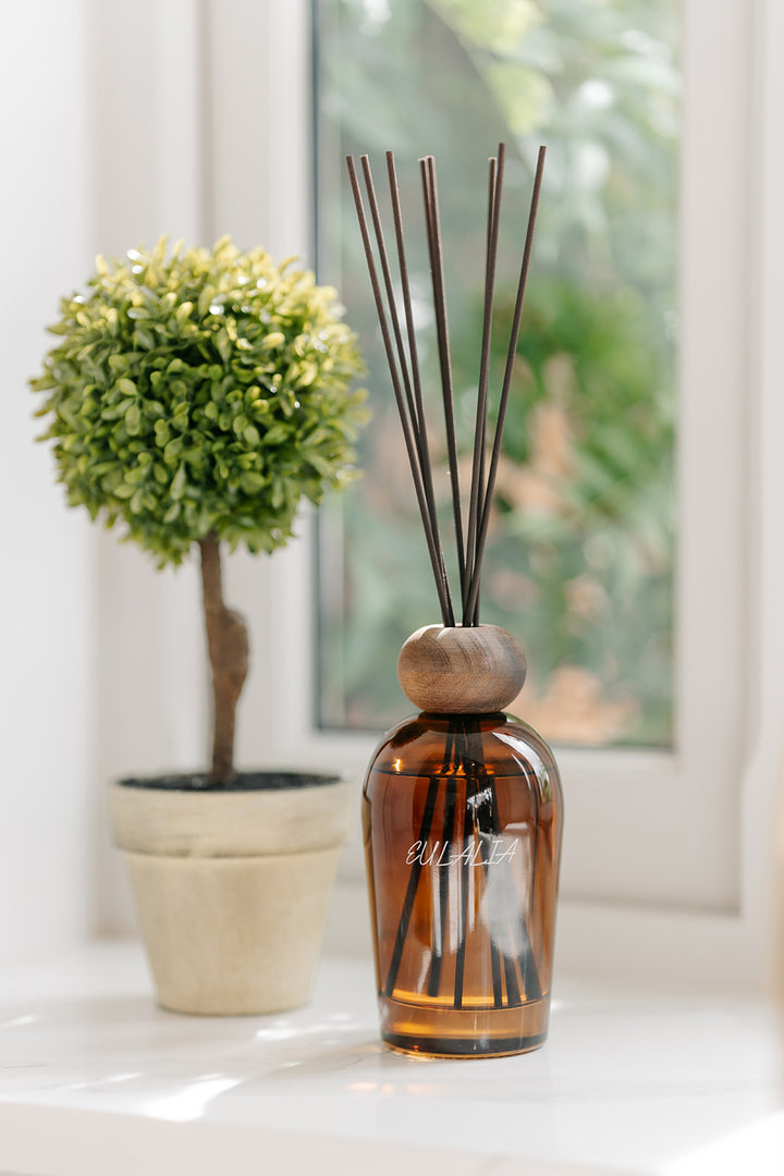 Amber glass reed diffuser bottle with a wooden stopper and dark reeds, placed on a white windowsill next to a potted green topiary tree.