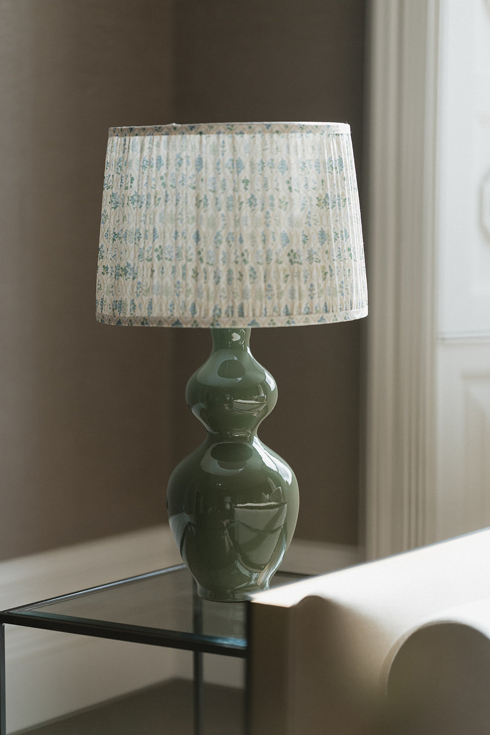 A table lamp with a glossy sage green double-bulb base and a pleated drum shade featuring a light blue botanical pattern, shown on a glass side table in a softly lit room.