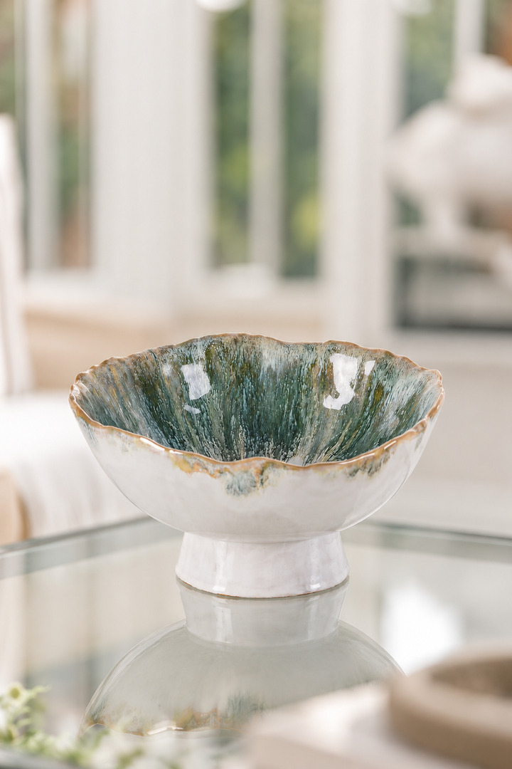 A ceramic pedestal bowl with a fluted rim, featuring a textured green and blue-green interior glaze and a rustic gold-brown edge, displayed on a glass table in a bright, modern room.