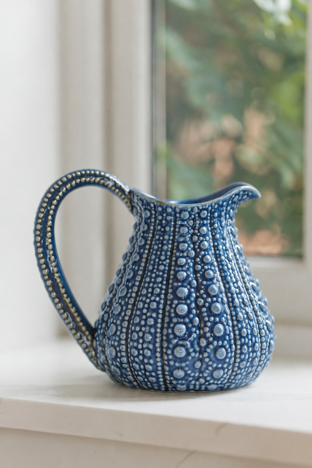 A blue ceramic pitcher with a textured, raised dot pattern and a curved handle, sitting on a white windowsill with a blurred window and greenery in the background.