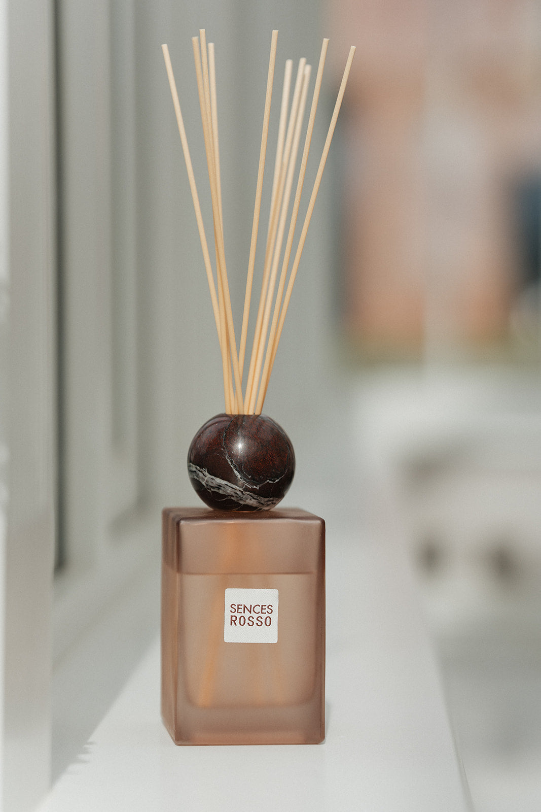 A reed diffuser bottle with a frosted amber-brown glass base, a dark polished stone sphere stopper, and natural reed sticks, shown on a white surface with a softly blurred indoor background. The front label reads &