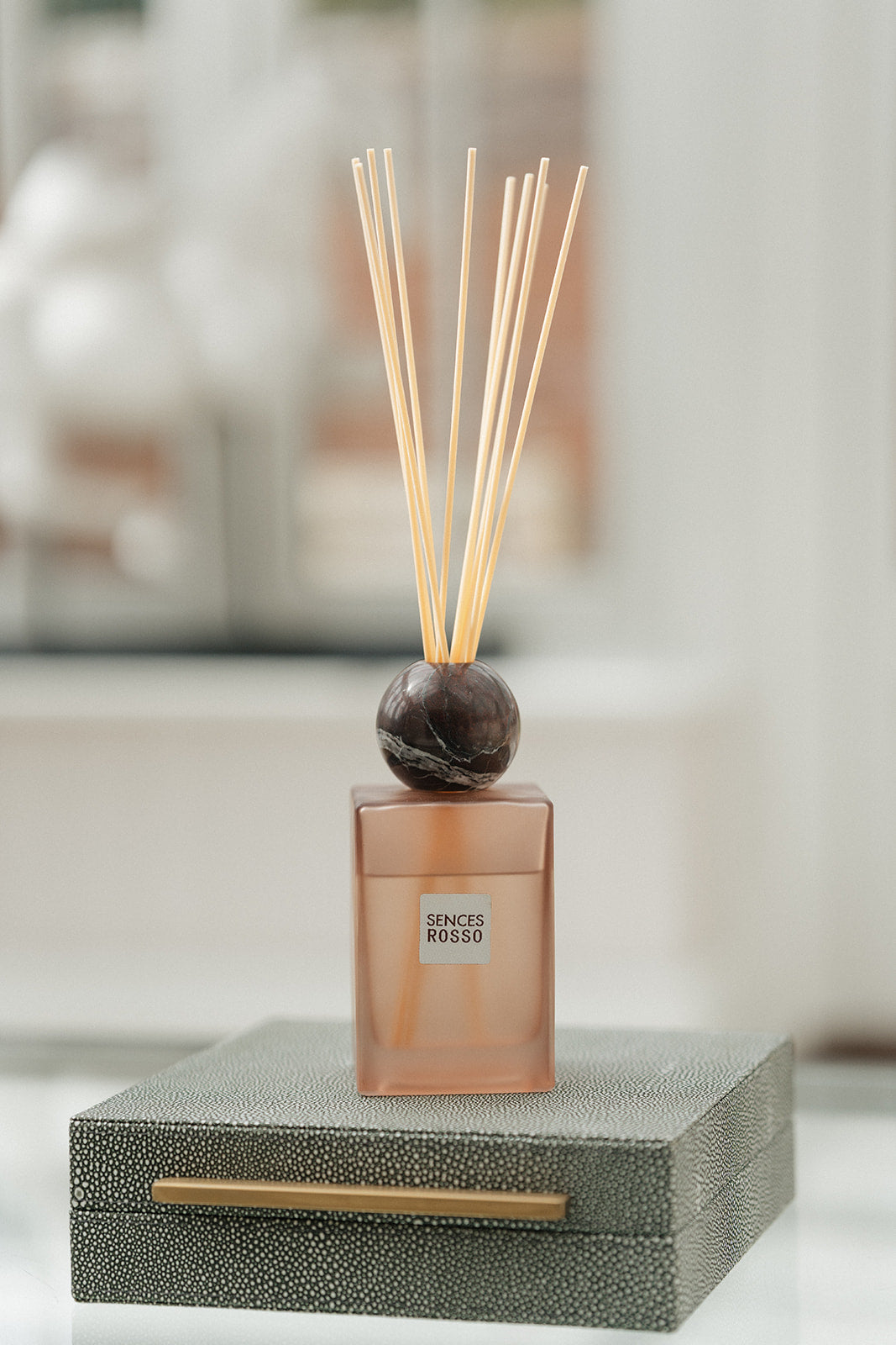 A reed diffuser bottle with a frosted amber-brown glass body, a dark round stopper, and natural reed sticks, displayed on top of a textured gray box with a gold handle.