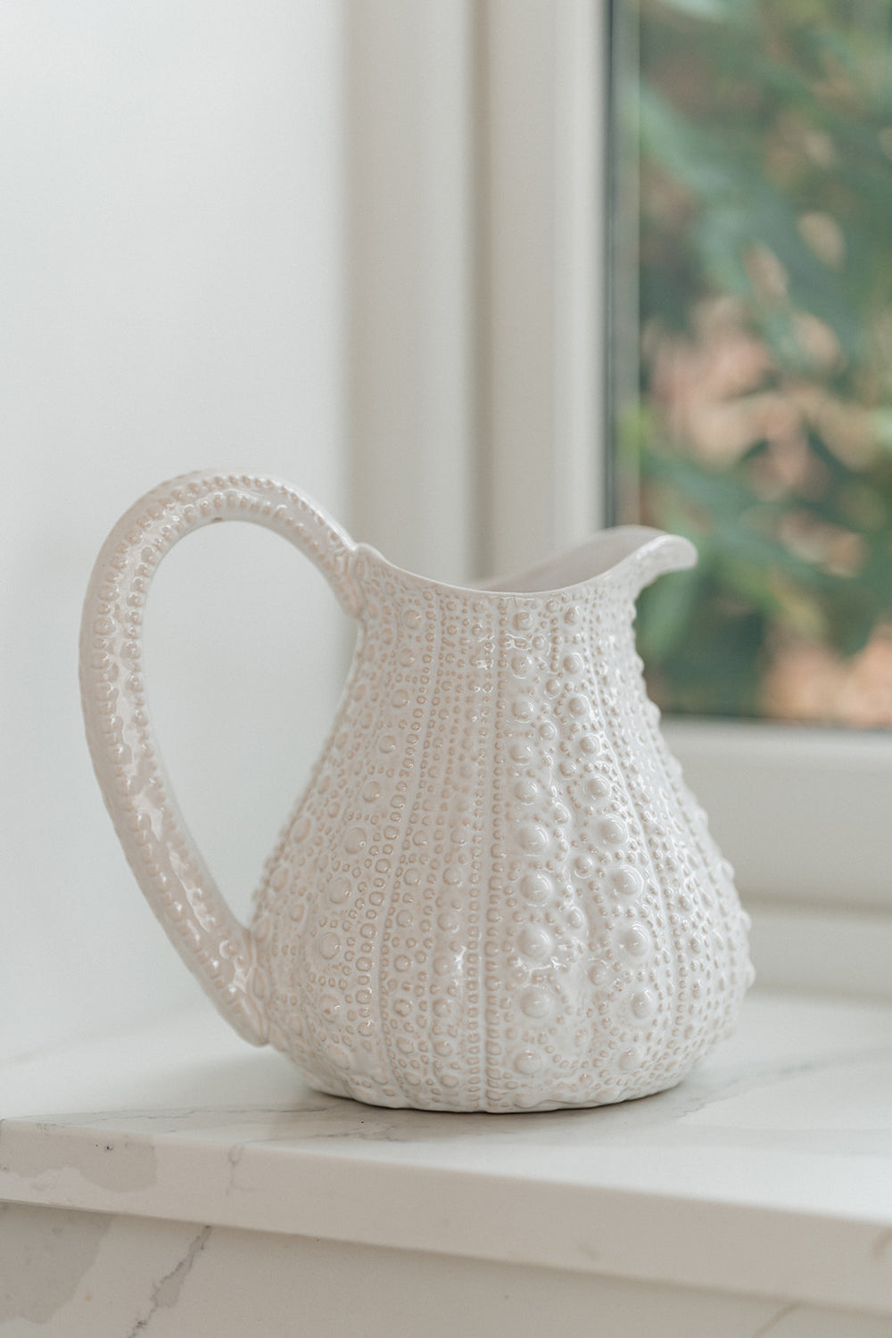 A white textured ceramic pitcher with a ribbed body and loop handle, resting on a white marble windowsill with a soft-focus window view in the background.