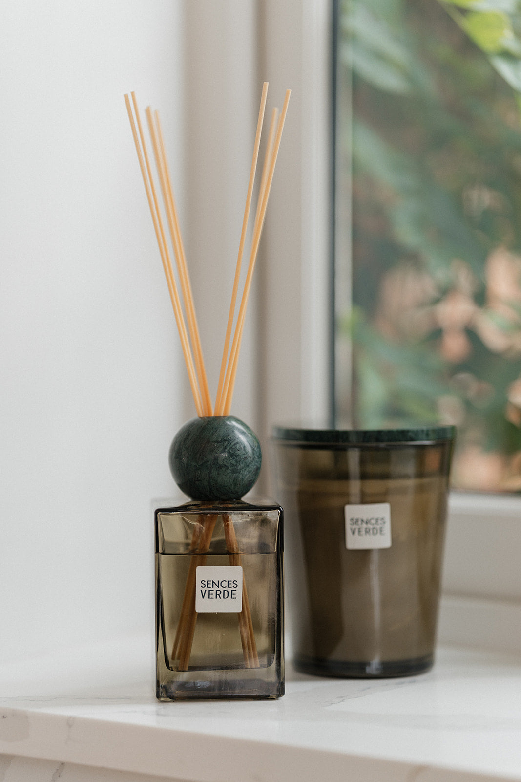 A reed diffuser bottle with a dark green spherical top and light wood reeds stands on a white windowsill next to a matching amber glass candle jar with a white label reading &