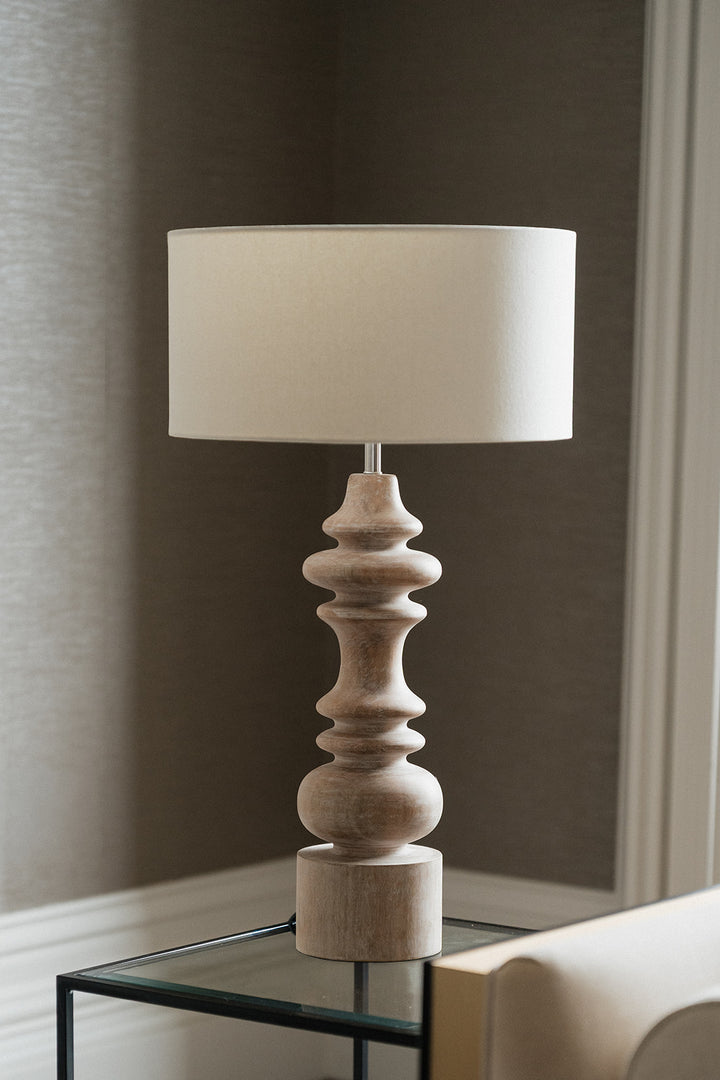 A table lamp with a cylindrical off-white fabric shade and a sculptural, multi-tiered turned wood base in a light beige finish, shown on a glass-topped side table against a neutral gray wall.