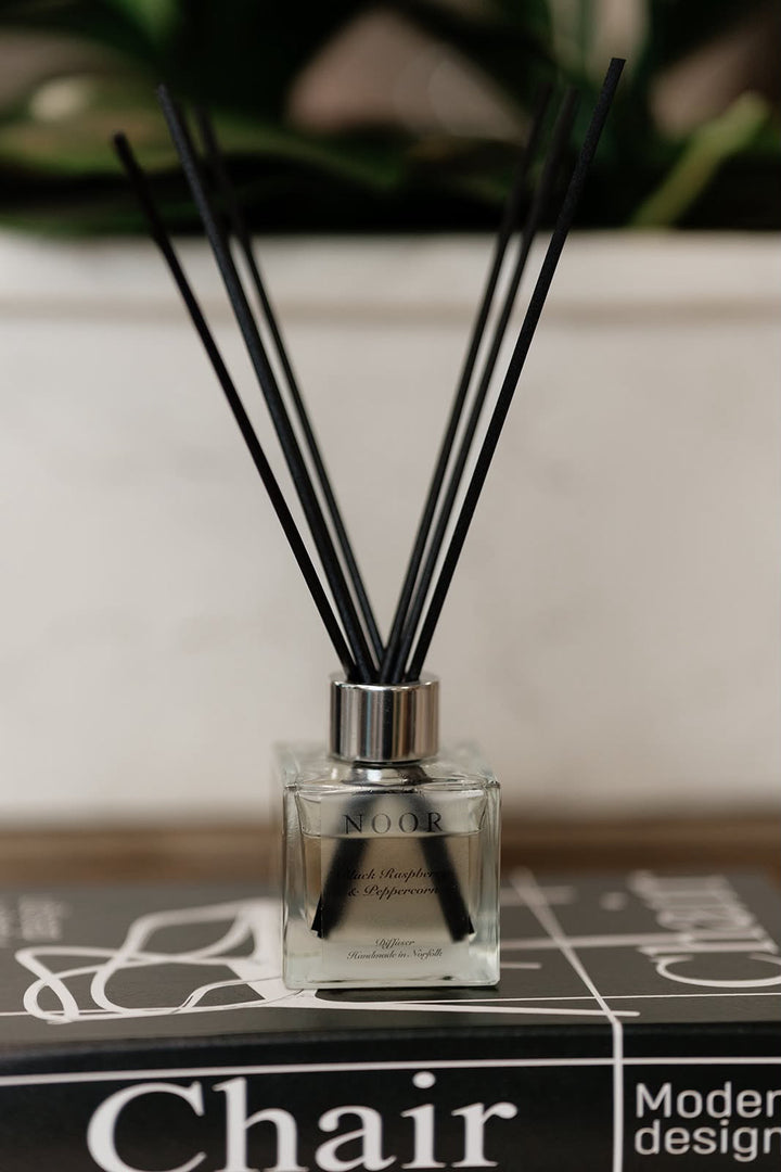 A clear glass reed diffuser bottle with black diffuser sticks, placed on top of a black book with the word &
