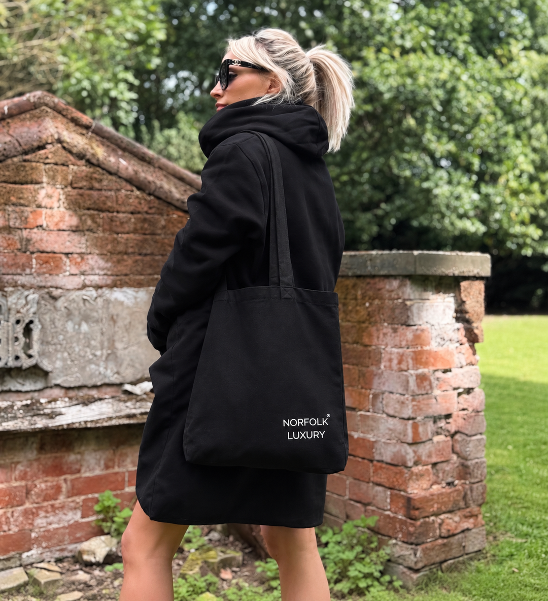 A woman wearing a black oversized hoodie and sunglasses is shown from the back, carrying a black canvas tote bag with white 'NORFOLK LUXURY' branding. She is standing outdoors in front of a brick wall and green trees.