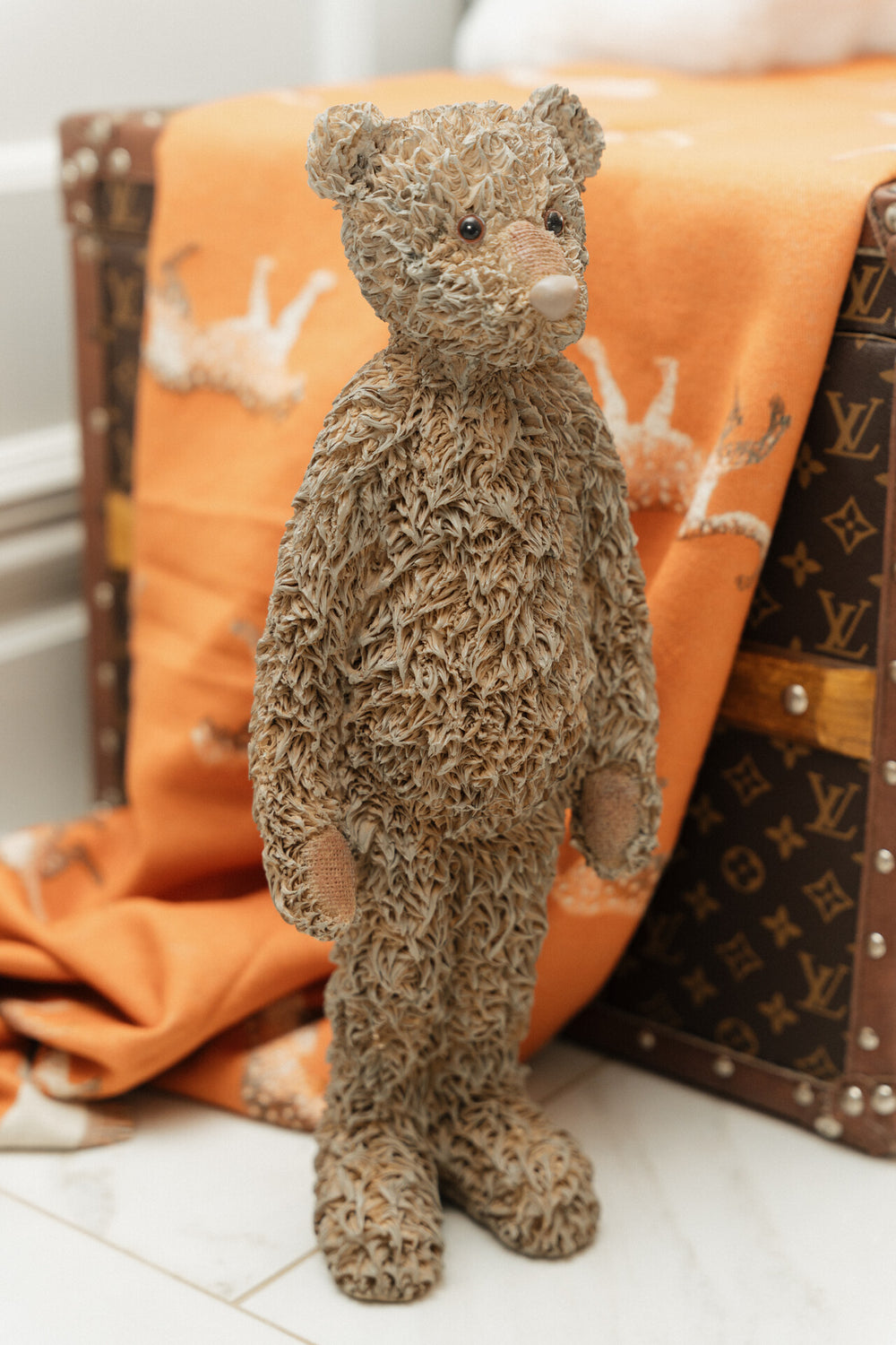 Brown teddy bear standing next to a patterned suitcase and orange blanket.