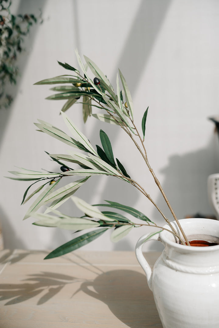Artificial olive branch stems with green and silvery-green leaves and small dark berries, arranged in a white ceramic pitcher on a light wooden surface against a white wall.