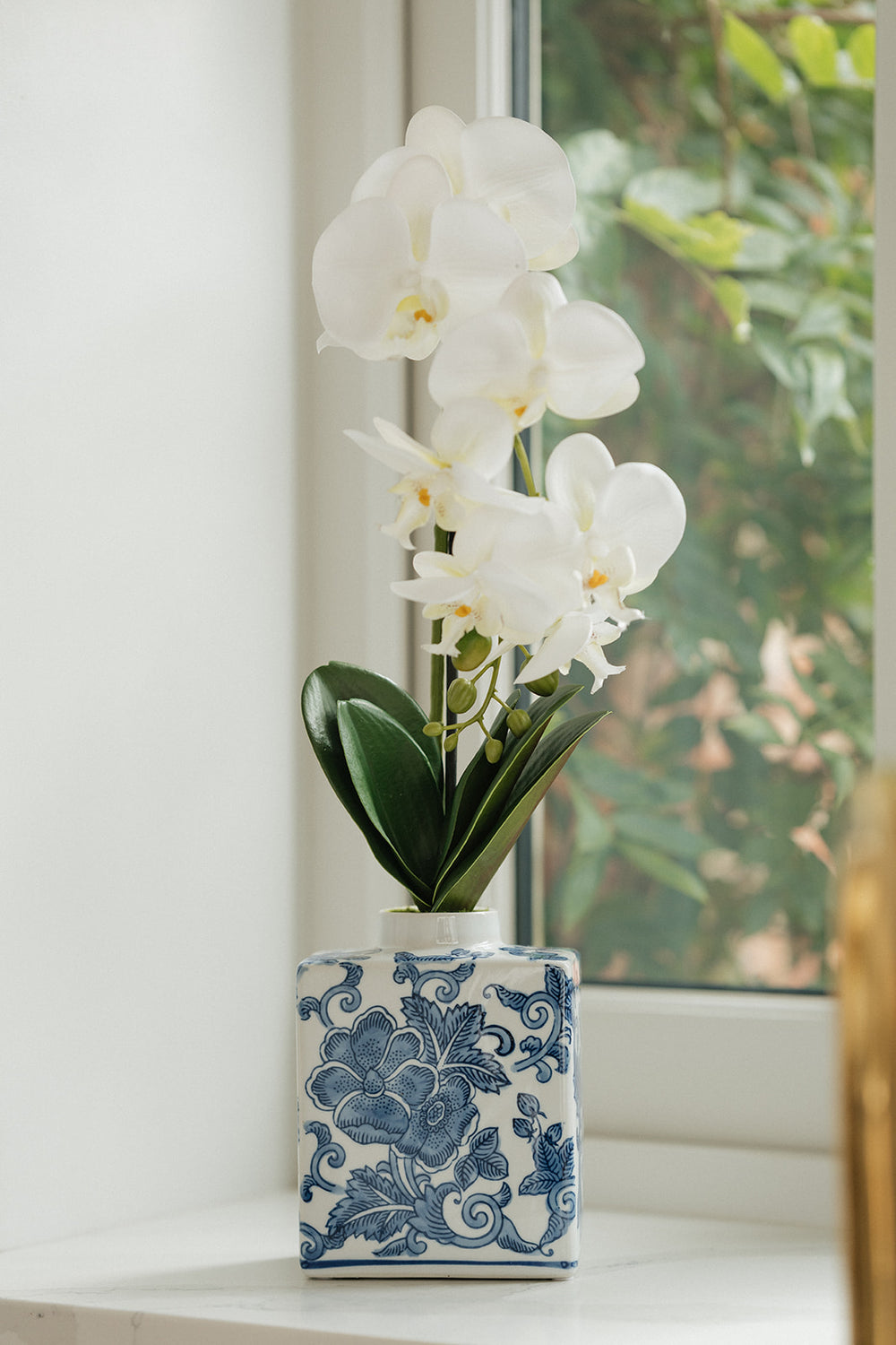 White artificial orchid flowers with green leaves arranged in a blue and white patterned ceramic vase, set on a windowsill with a blurred window and greenery in the background.