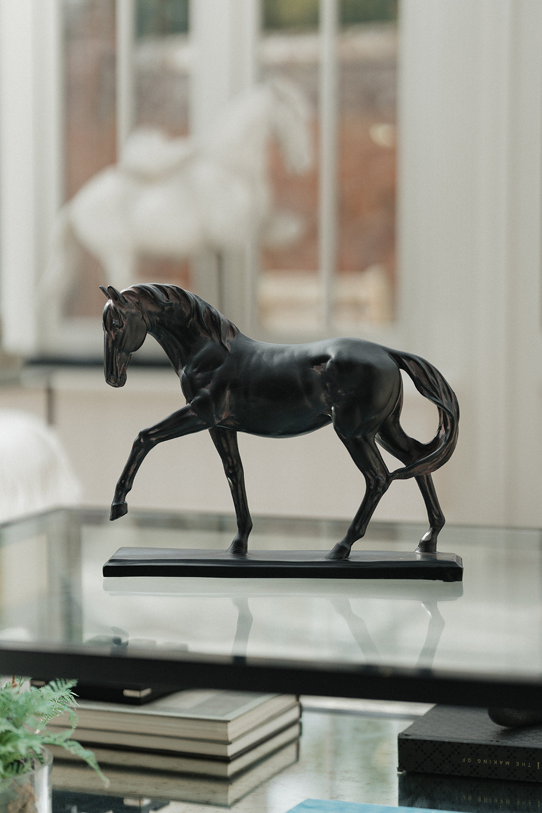 A dark bronze horse figurine standing on a rectangular base, displayed on a glass tabletop in a softly lit interior with blurred books and a window in the background.