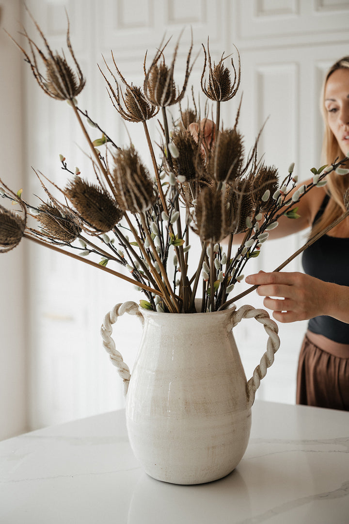 A woman arranging dried botanical stems in a large white ceramic vase with rope handles, set on a light marble table against a white paneled wall.