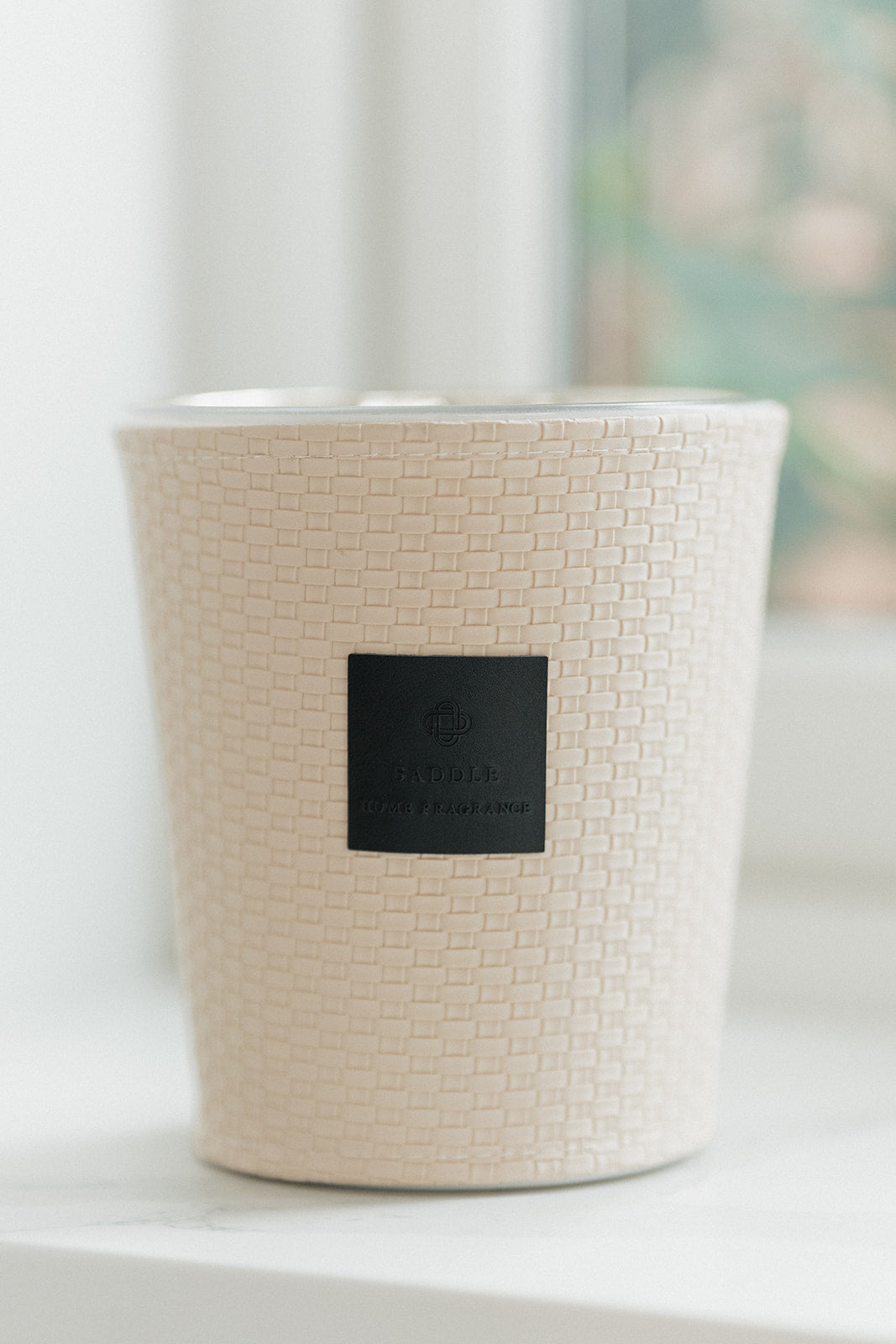 A textured cream-colored candle jar with a black square label in the center, resting on a white surface with a softly blurred window background.