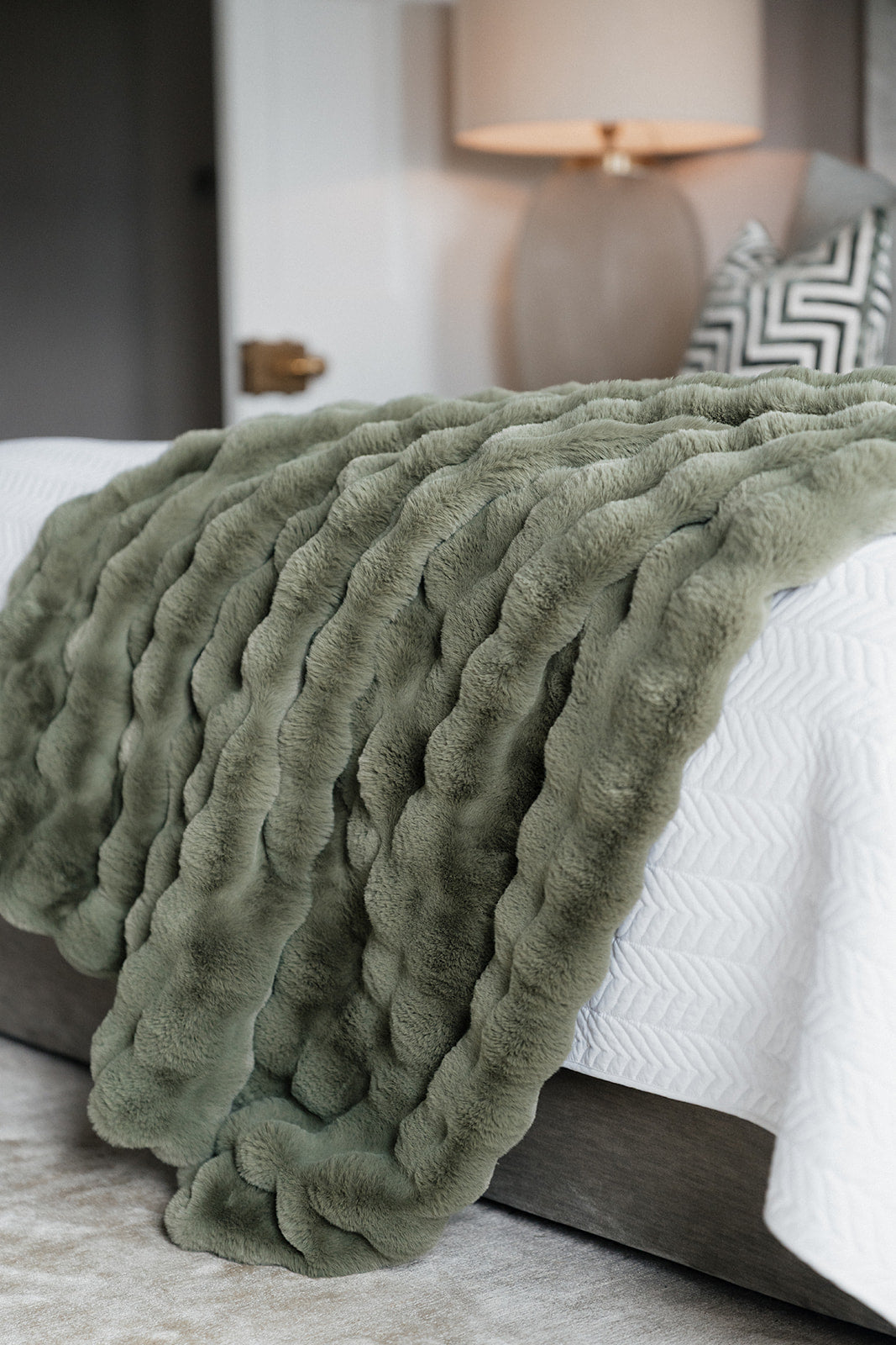 An olive green chunky faux fur throw blanket draped over a bed with white chevron bedding in a softly lit bedroom setting.
