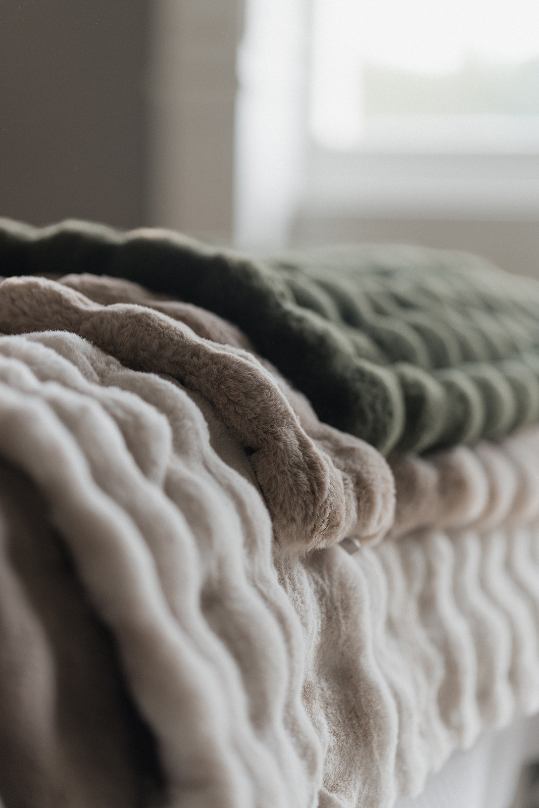 Three textured, ribbed knit blankets in cream, taupe, and olive green stacked on a bed in a softly lit bedroom.