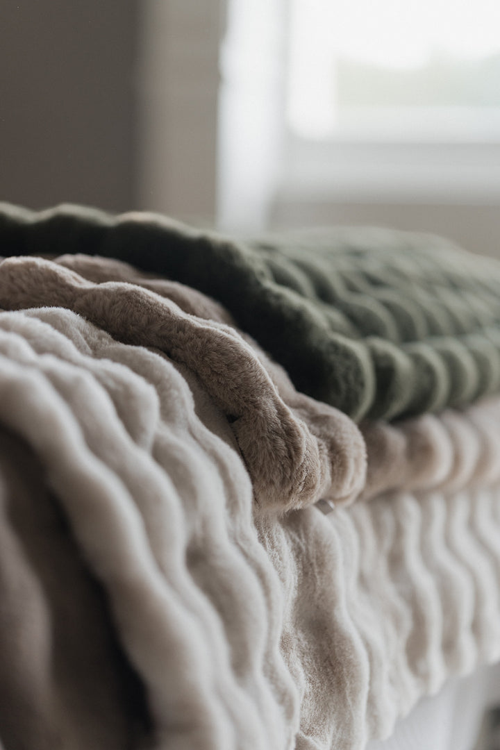 Three textured, ribbed knit blankets in cream, taupe, and olive green stacked on a bed in a softly lit bedroom.