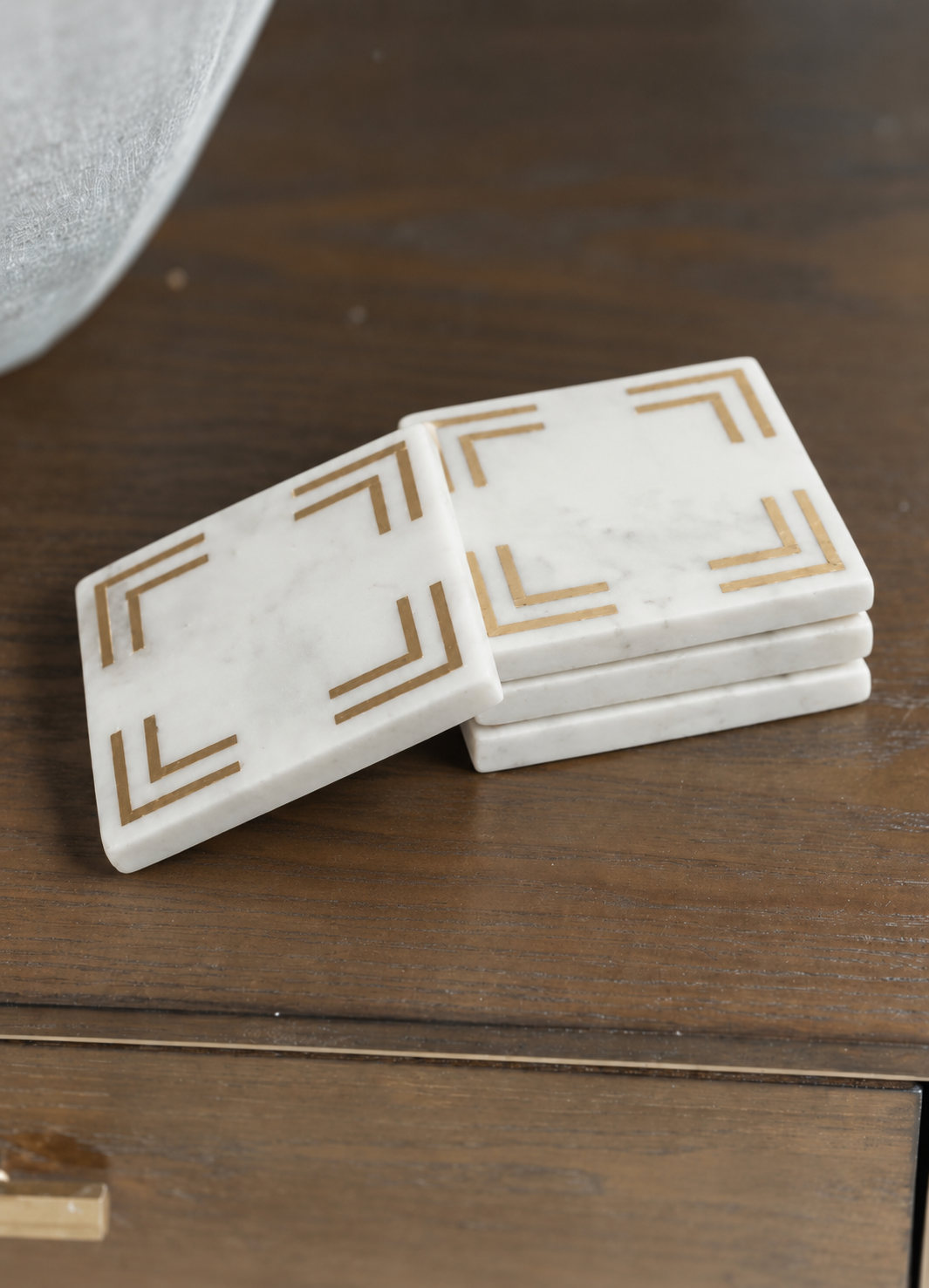 A stack of four square white marble coasters with gold geometric corner designs, resting on a dark wood table.