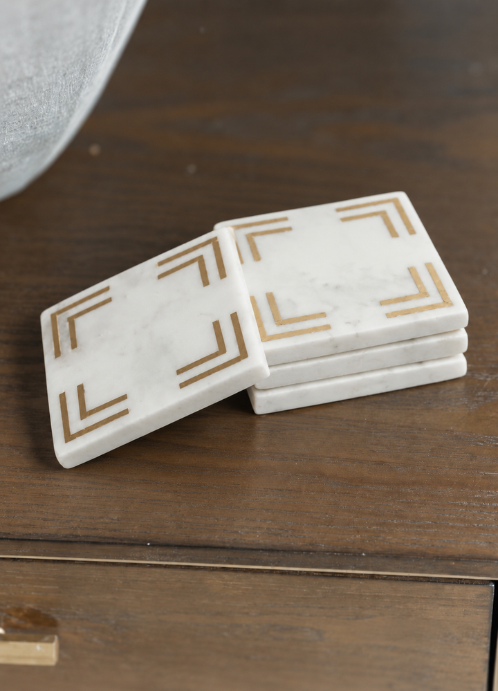 A stack of four square white marble coasters with gold geometric corner designs, resting on a dark wood table.