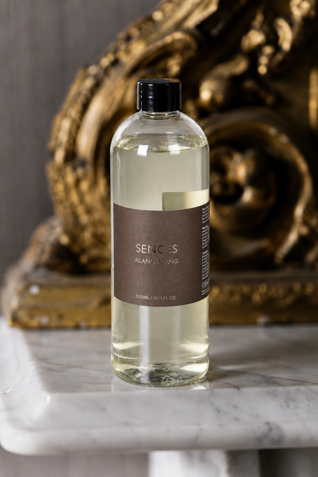A clear bottle of Senses Alan Puring cleansing oil with a black cap and brown label, set on a white marble surface with an ornate gold sculpture in the background.