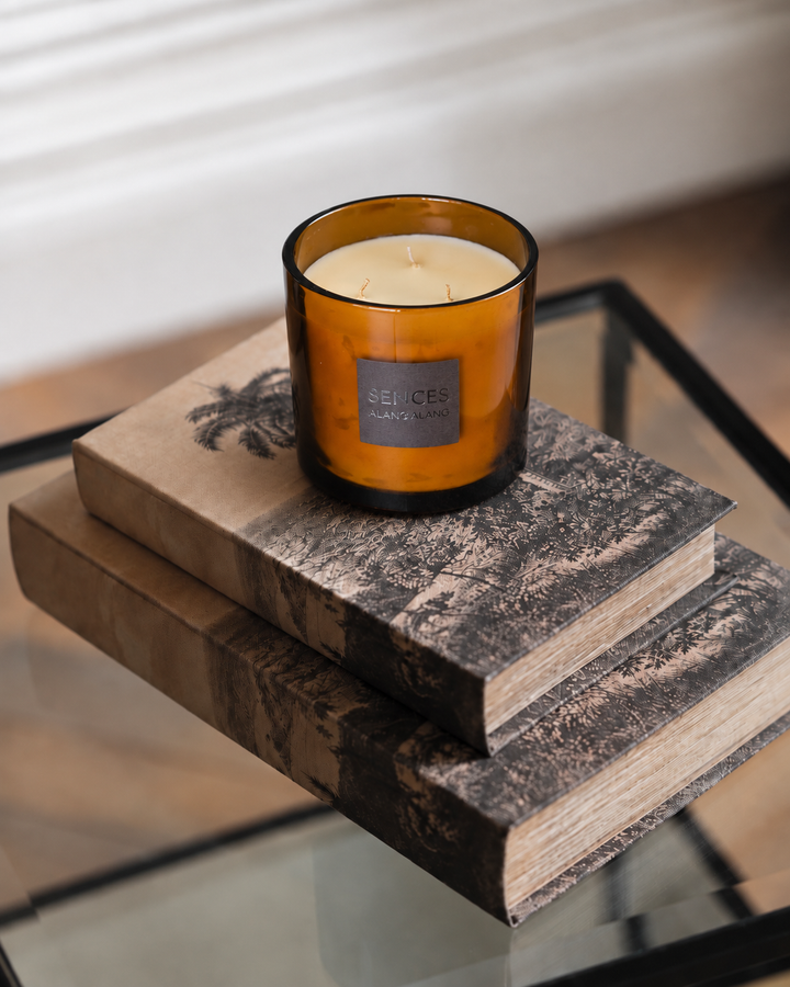 An amber glass jar candle with cream wax sits on a stack of two rustic books on a glass table. The candle label reads &