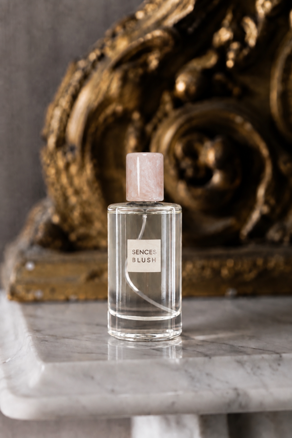 A clear rectangular perfume bottle with a pale pink cap stands on a white marble surface in front of an ornate gold sculpture. The bottle label reads 'SENSE BLUSH'.