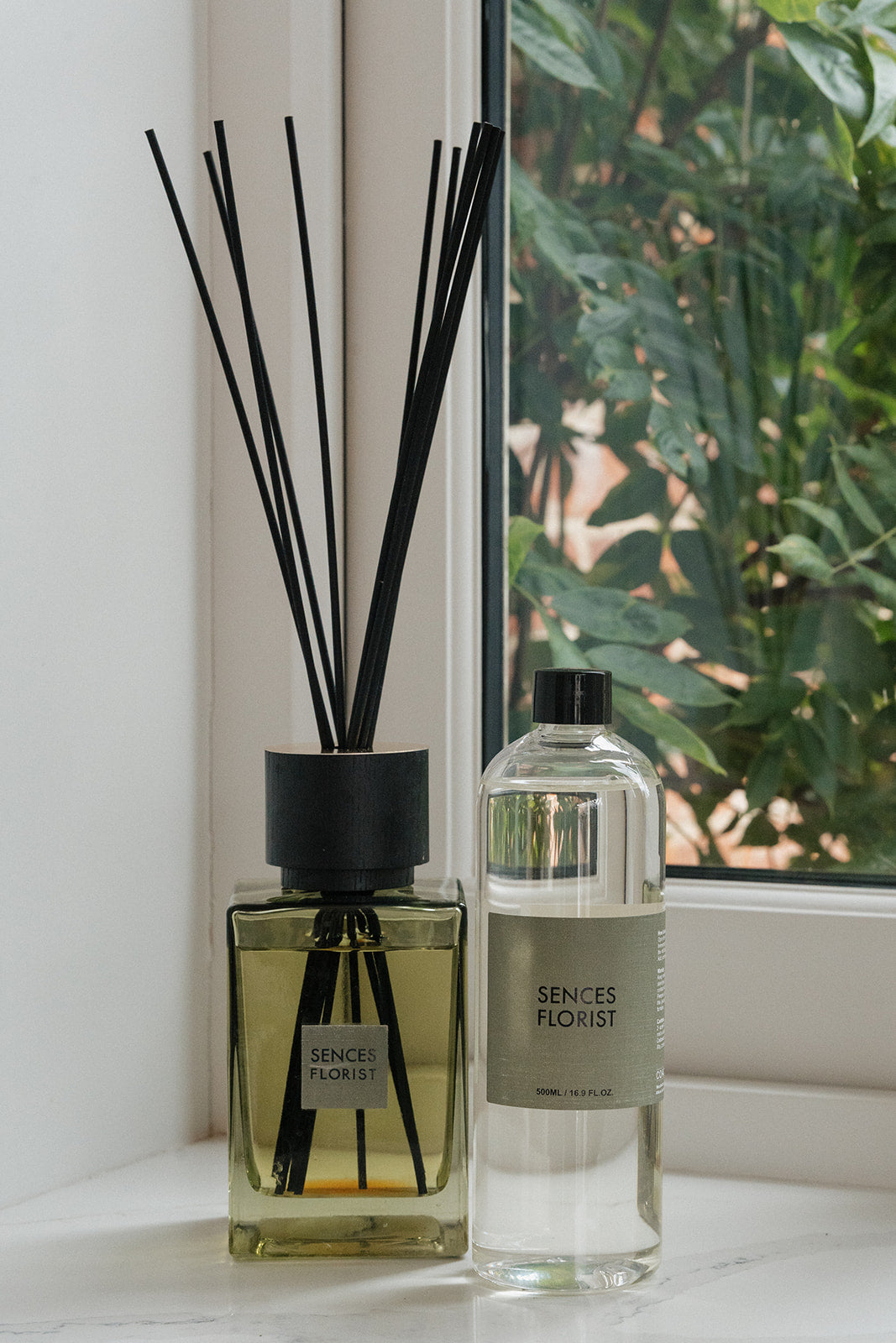 A reed diffuser bottle with black reeds and a clear bottle of liquid beside it, both labeled SENCES FLOHIST, set on a windowsill with a window view of green foliage in the background.