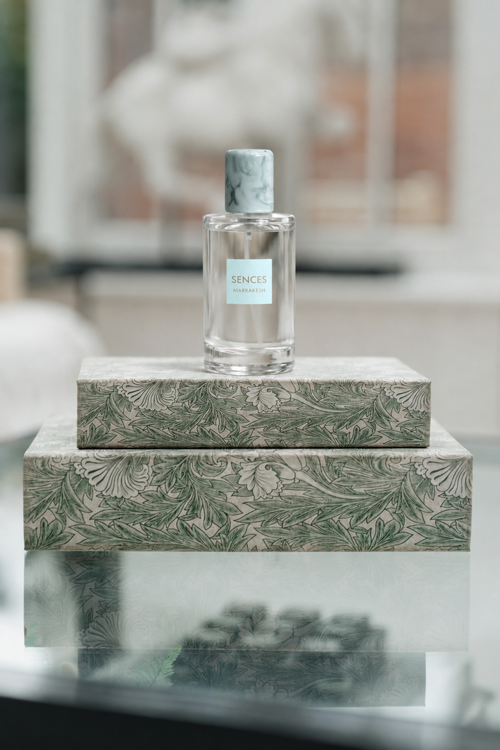 A clear glass perfume bottle with a light blue label and a gray-green marble cap sits centered on top of two patterned rectangular boxes with a green botanical print on a beige background. The scene is softly lit and photographed in a real interior setting with blurred background elements.